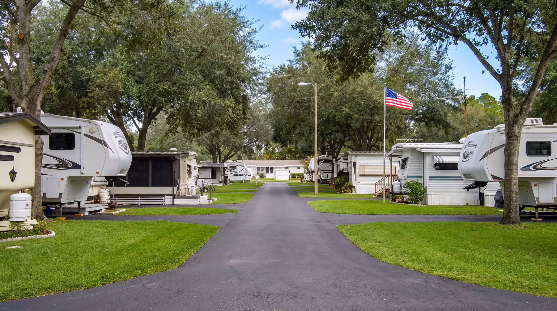 Paved lane through an RV park lined with campers under mossy oaks, American flag waving overhead.