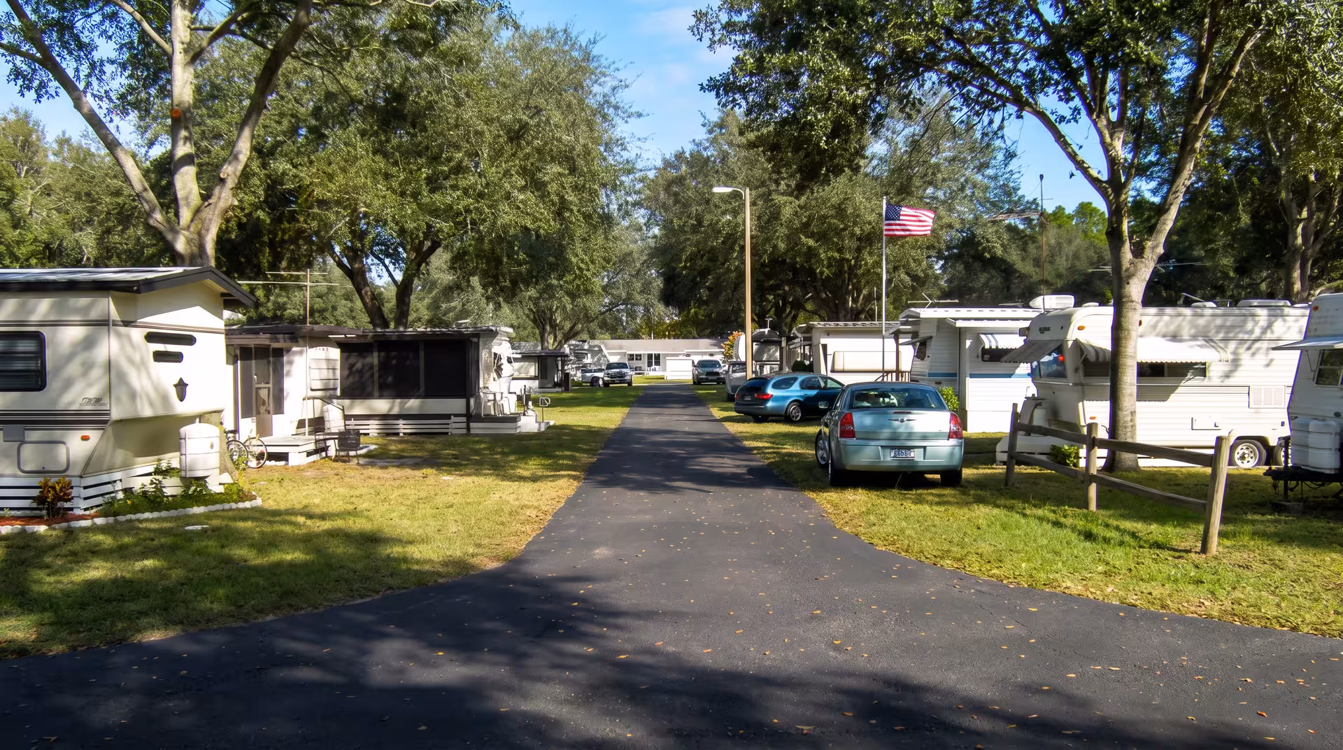 Paved road through RV park with campers, large shade trees, parked cars, and American flag.