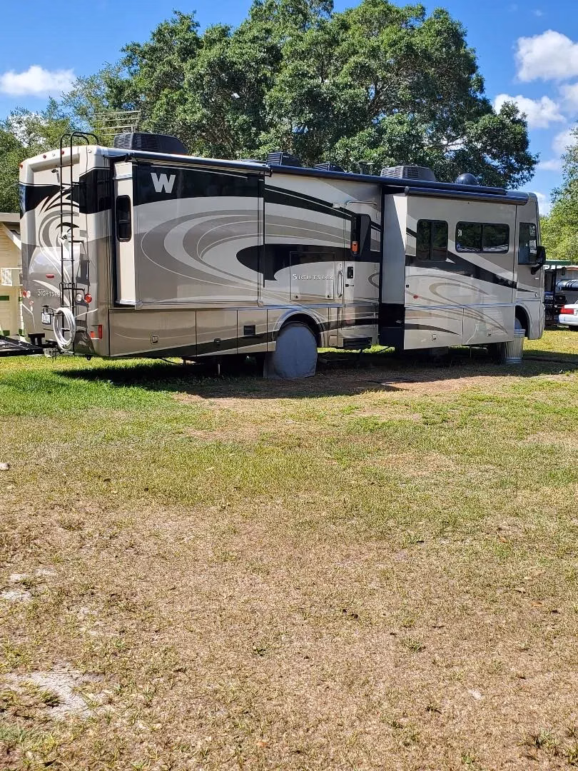 Winnebago motorhome with slide-outs extended, parked on a grassy RV site beneath shady trees